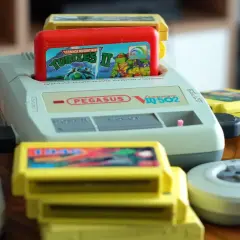 Konsola Pegasus IQ-502 z włożoną grą Teenage Mutant Ninja Turtles II i kilkoma żółtymi kartridżami oraz dwoma szarymi kontrolerami.