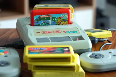 Konsola Pegasus IQ-502 z włożoną grą Teenage Mutant Ninja Turtles II i kilkoma żółtymi kartridżami oraz dwoma szarymi kontrolerami.