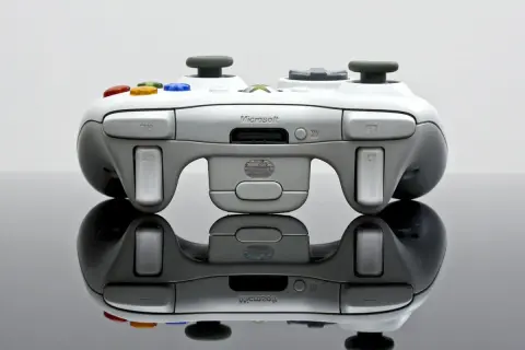 Biały kontroler Xbox 360 z kolorowymi przyciskami, odbijający się od czarnej powierzchni.