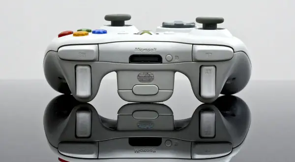 Biały kontroler Xbox 360 z kolorowymi przyciskami, odbijający się od czarnej powierzchni.