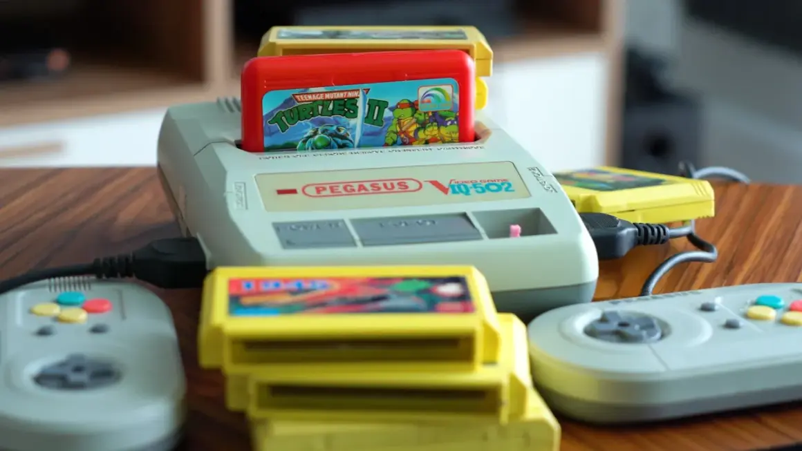 Konsola Pegasus IQ-502 z włożoną grą Teenage Mutant Ninja Turtles II i kilkoma żółtymi kartridżami oraz dwoma szarymi kontrolerami.