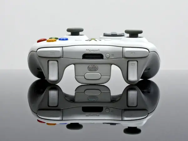Biały kontroler Xbox 360 z kolorowymi przyciskami, odbijający się od czarnej powierzchni.